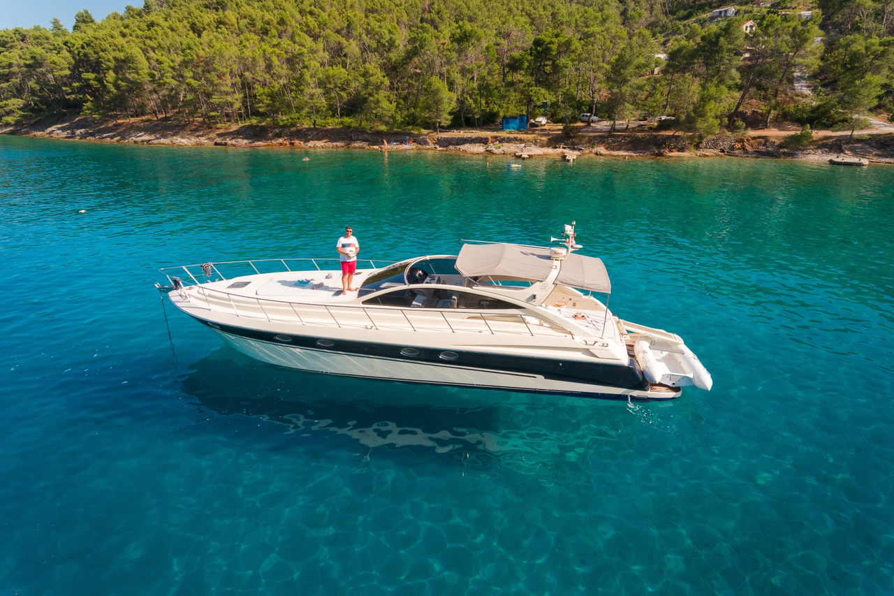 Aerial view of a crewed motor yacht anchored over sand in a pine-fringed Dalmatian bay near Split, with a guest standing on the bow.