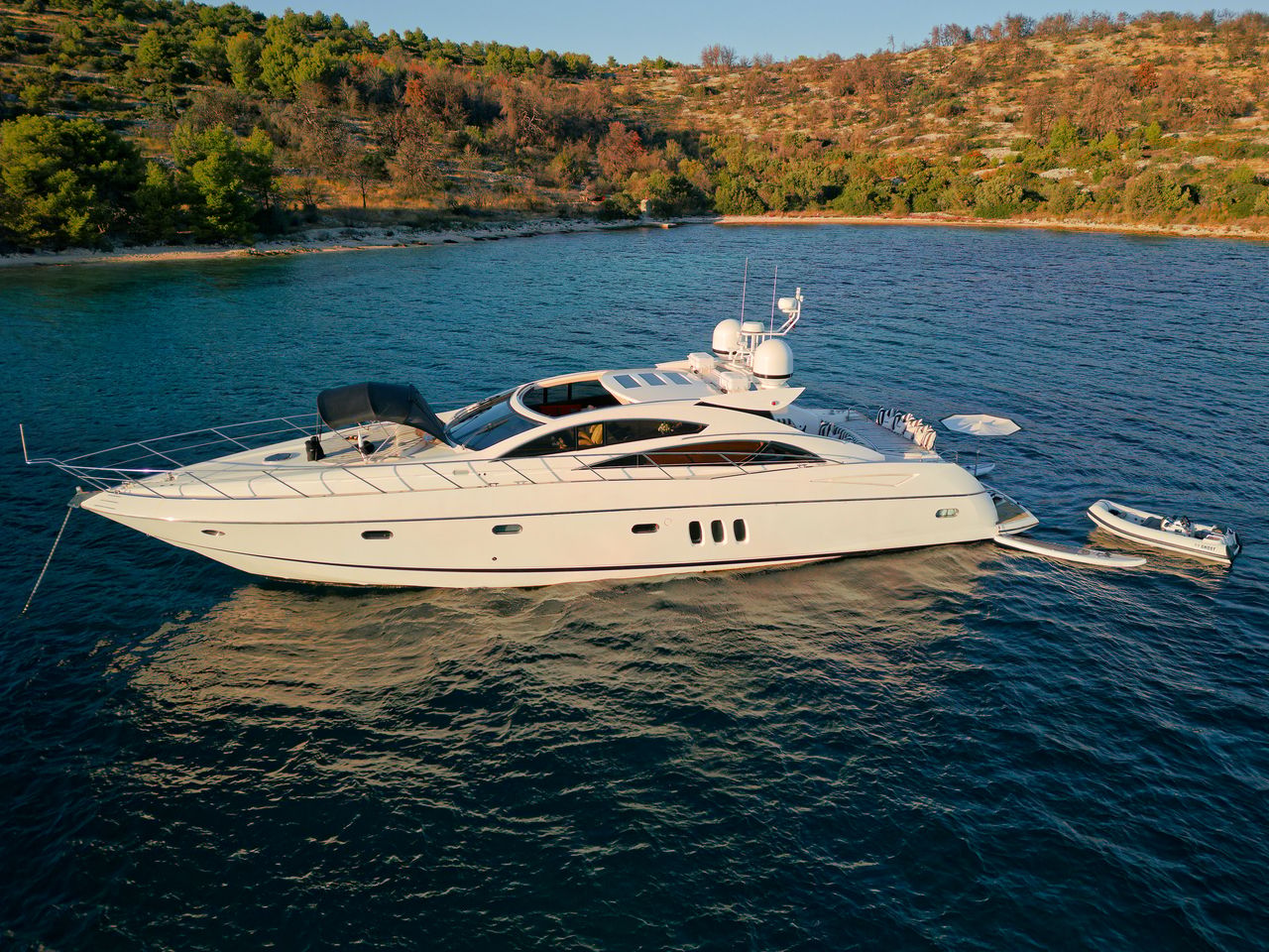 Aerial side view of a crewed motor yacht at anchor off a wooded Dalmatian island, tender made fast astern, in late afternoon light.