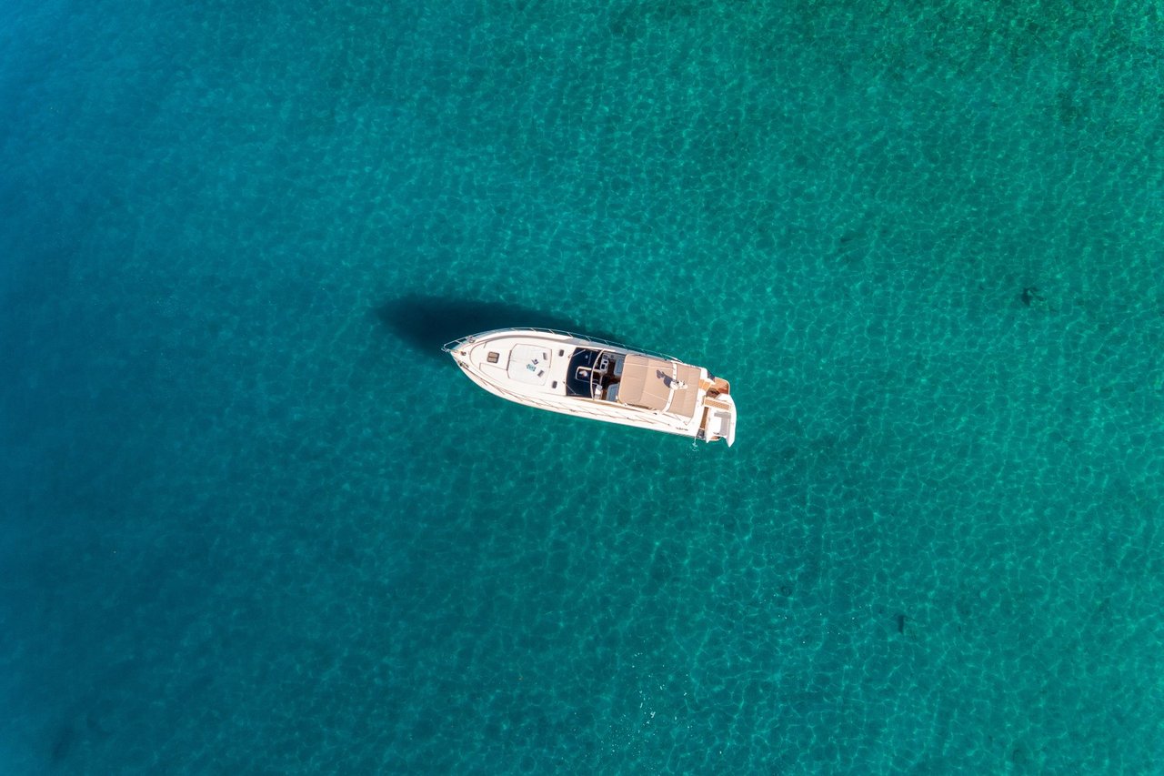 Aerial view of a small motor yacht lying at anchor over pale sand in clear turquoise water, its shadow visible on the seabed, off the Dalmatian coast.
